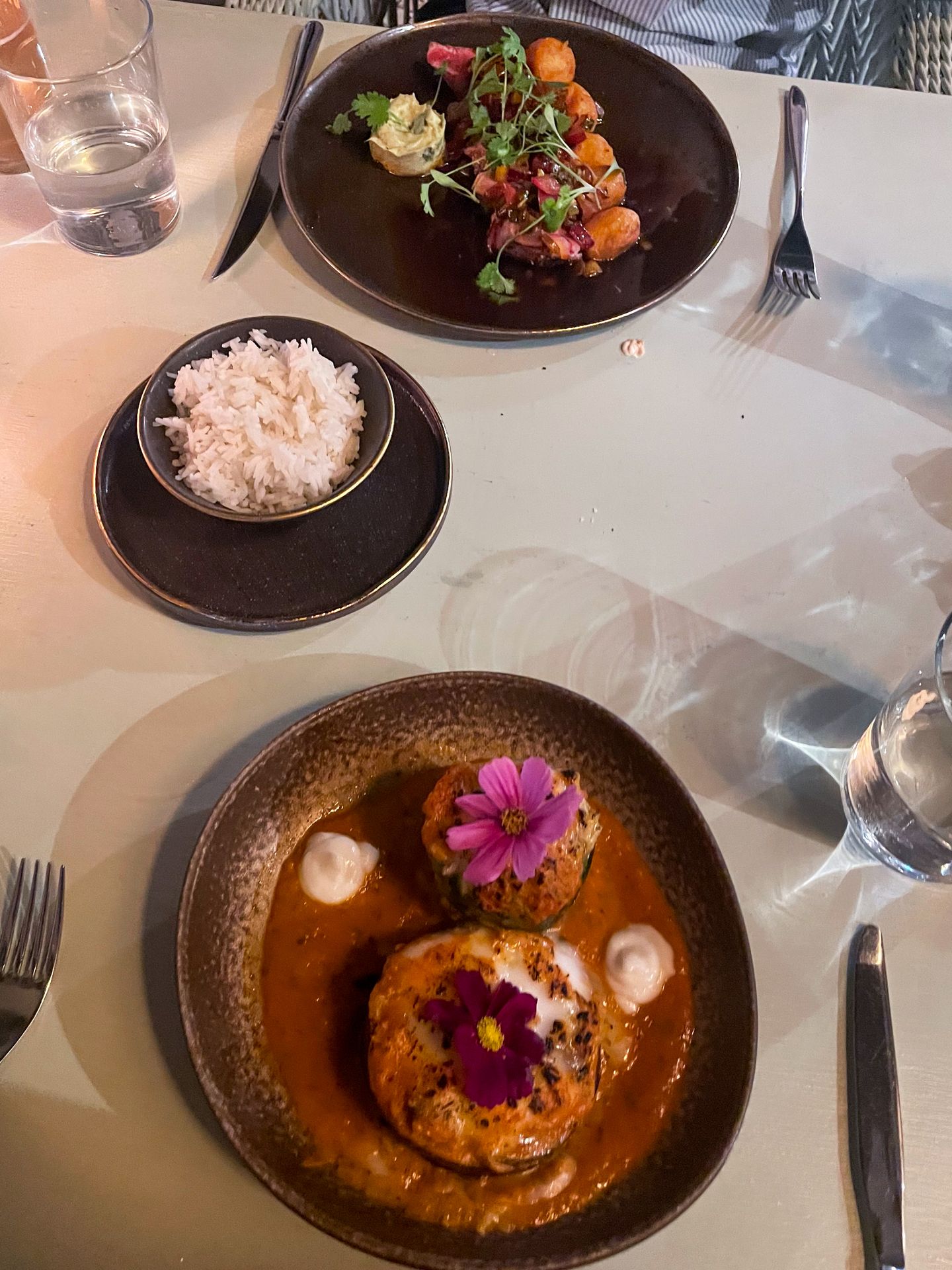 A plate with two round veggies covered in cheese, topped with flowers and surrounded by sauce. There is also rice and a meat dish on the table.