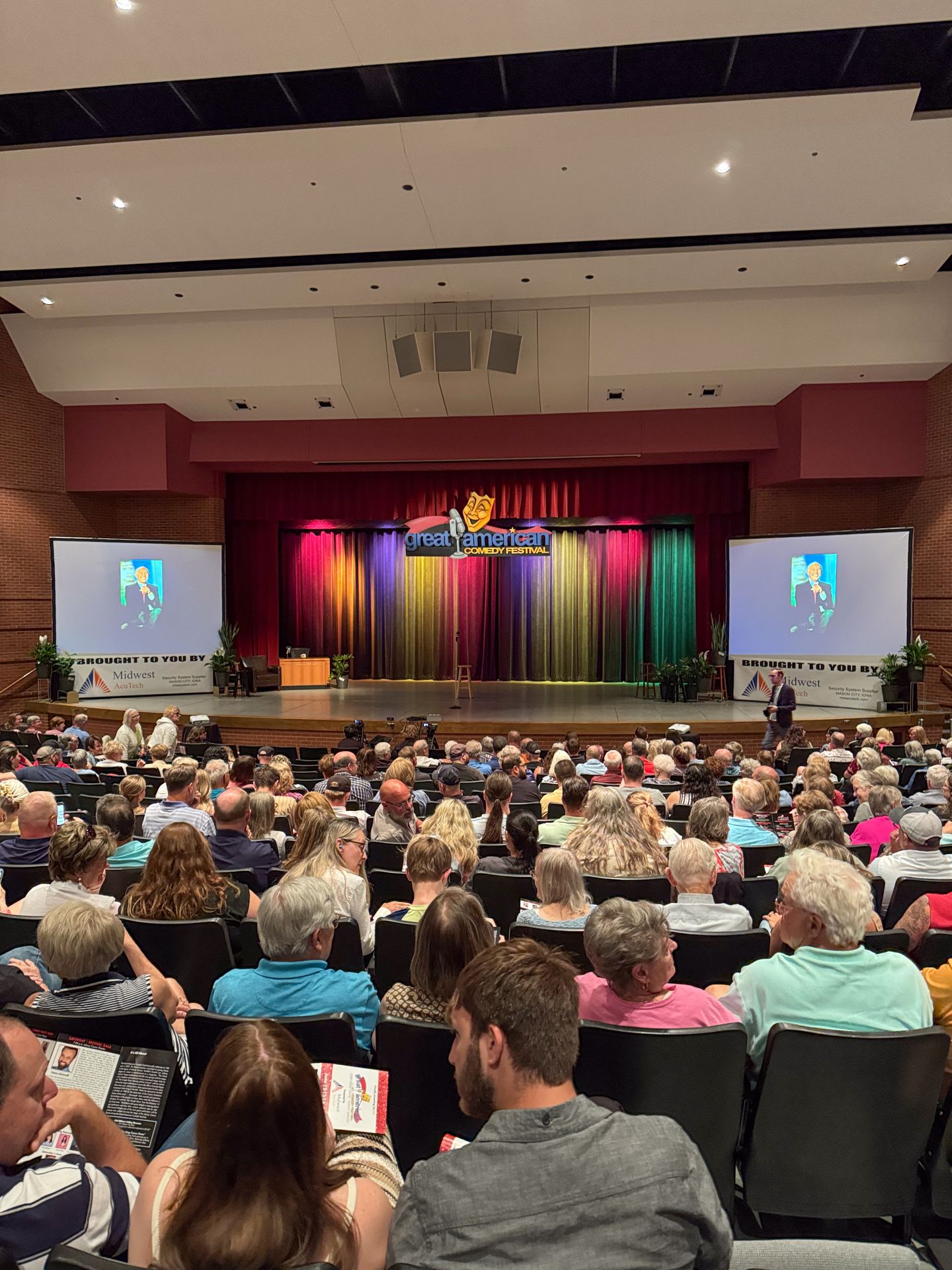Looking down at a stage with a full audience inside the Johnny Carson Theatre. The stage curtains are lit with rainbow colors