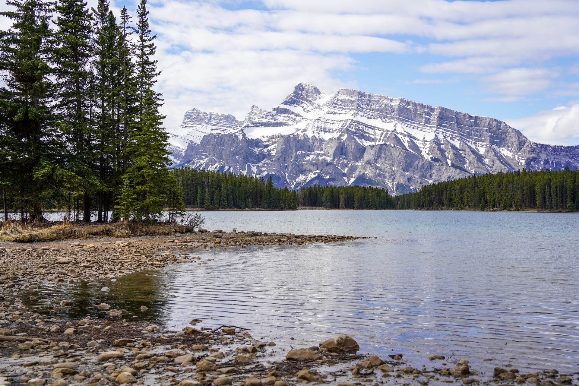 A rocky beach and a little peninsula. Across the water is a mountain partially covered in snow.
