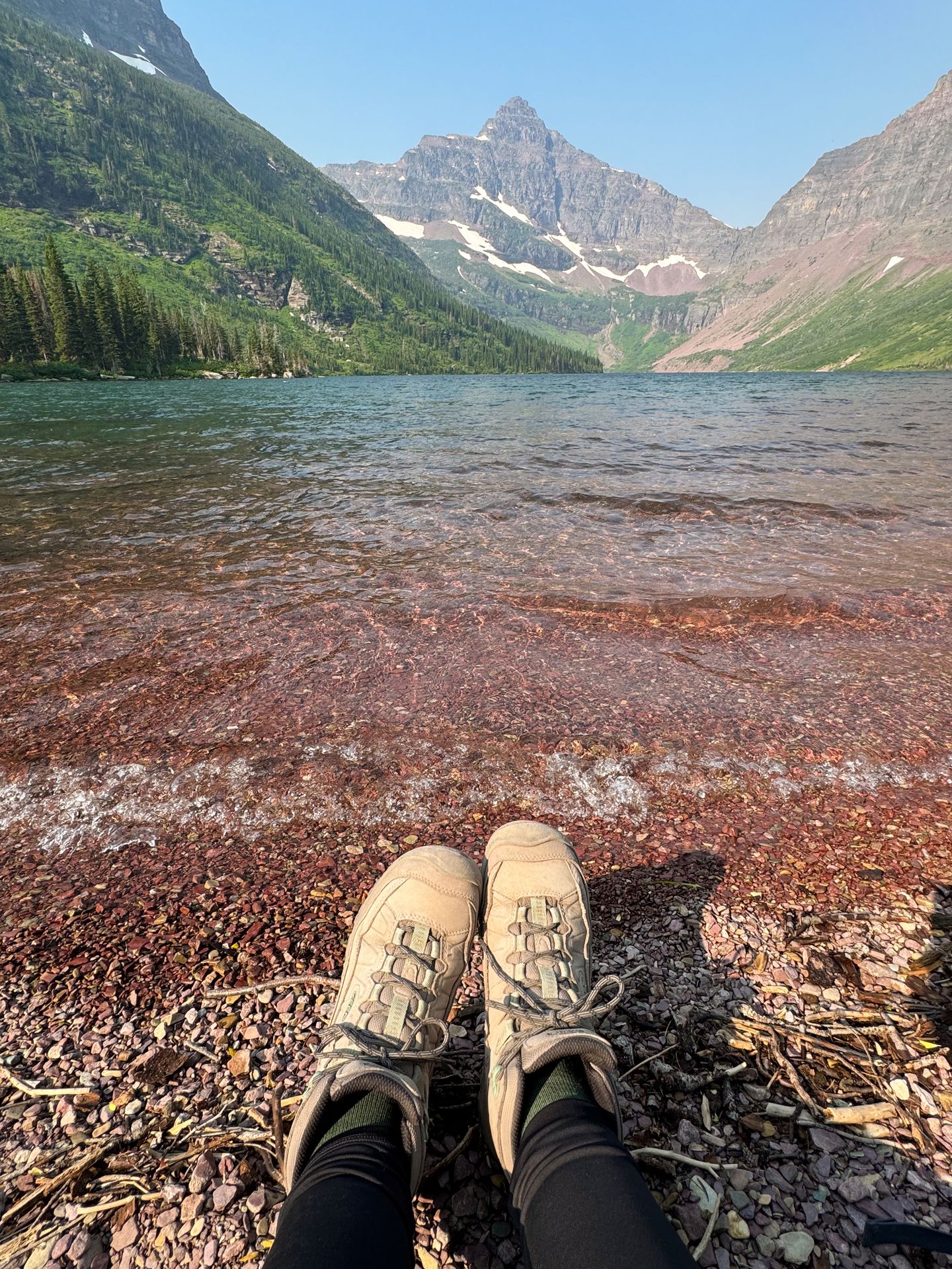 Looking at a pair of feet on the edge of Upper Two Medicine Lake. The nearby rocks have a reddish color.