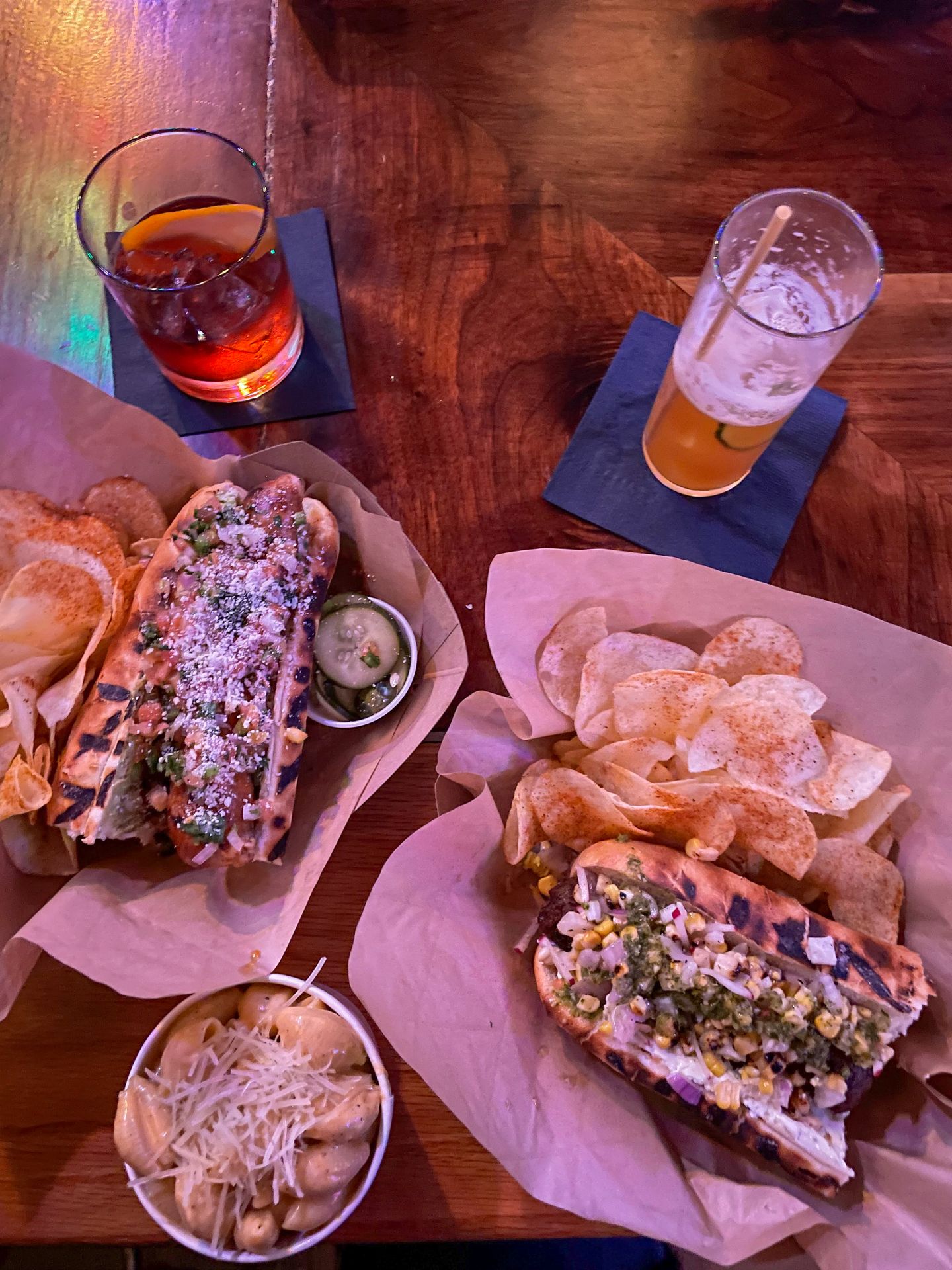 Two hot dogs made with grilled bread, served with chips. There is also a cup of mac and cheese and two cocktails on the table.