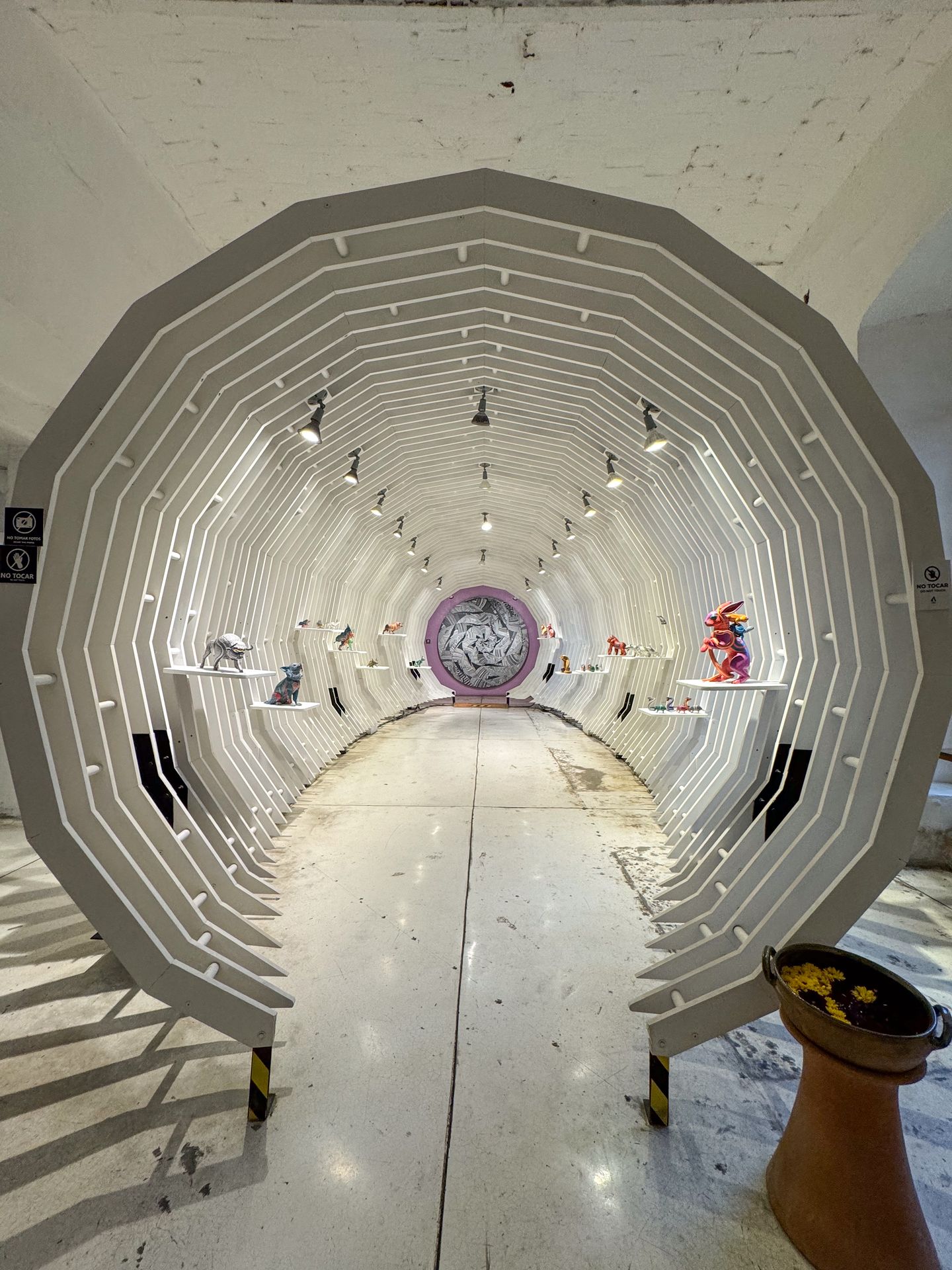 A white, skeletal-feeling tunnel with sculptures hanging on sheleves at Voces de Copal