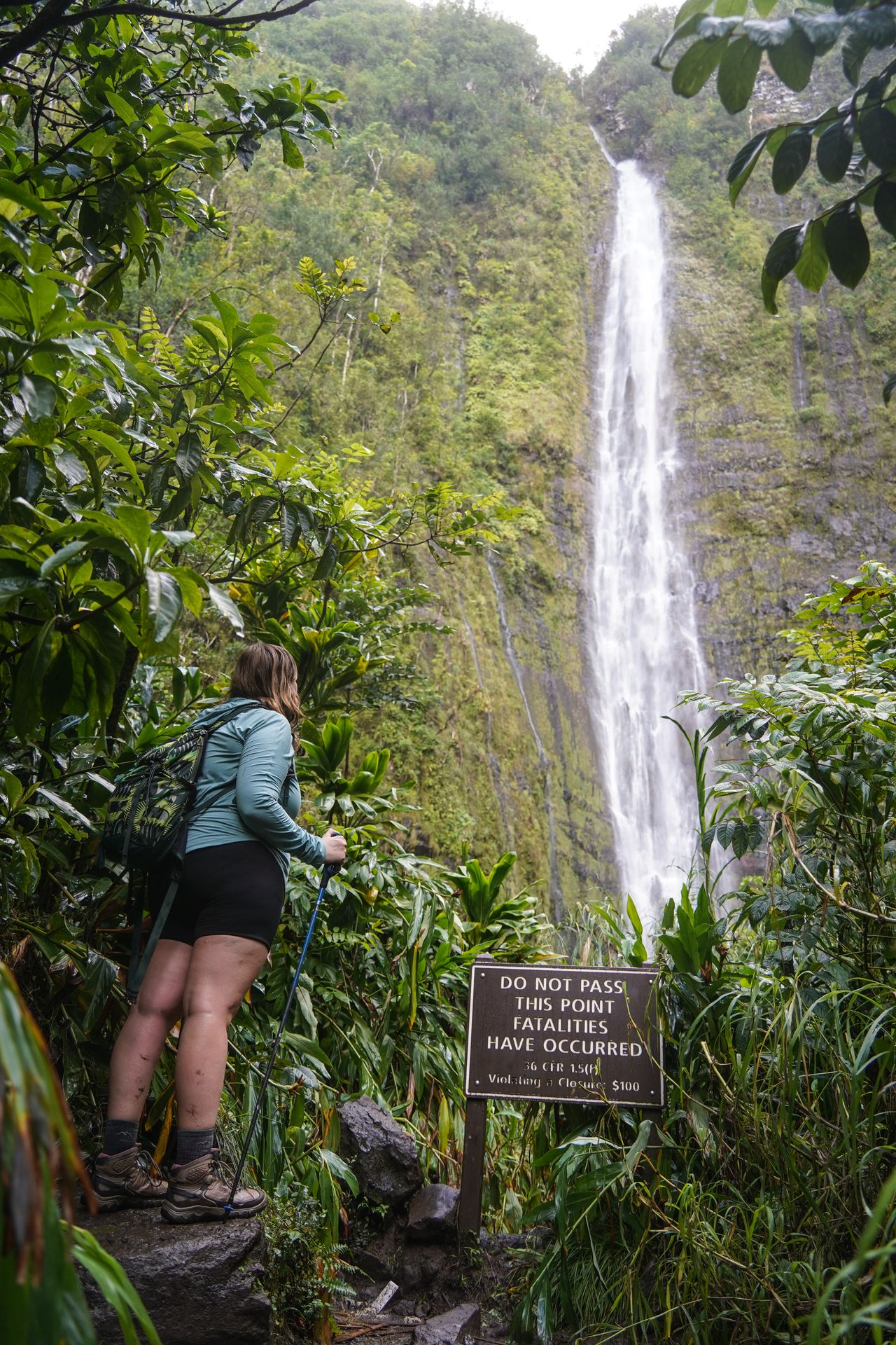 Lydia looking up at a tall waterfall. A sign reads 'Do not cross this point fatalaties have occured'