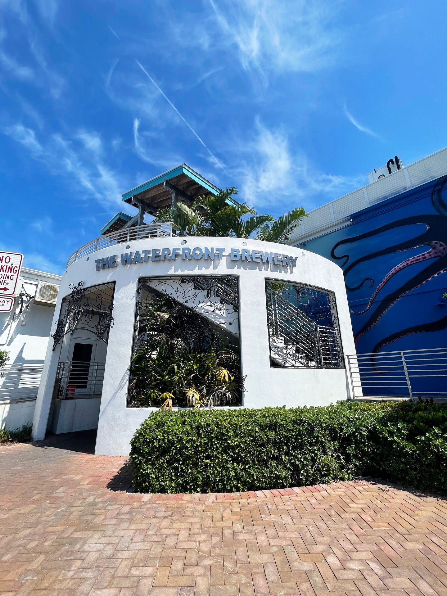 An exterior view of a white round building labeled The Waterfront Brewery. Greenery is coming out of and on top of the building