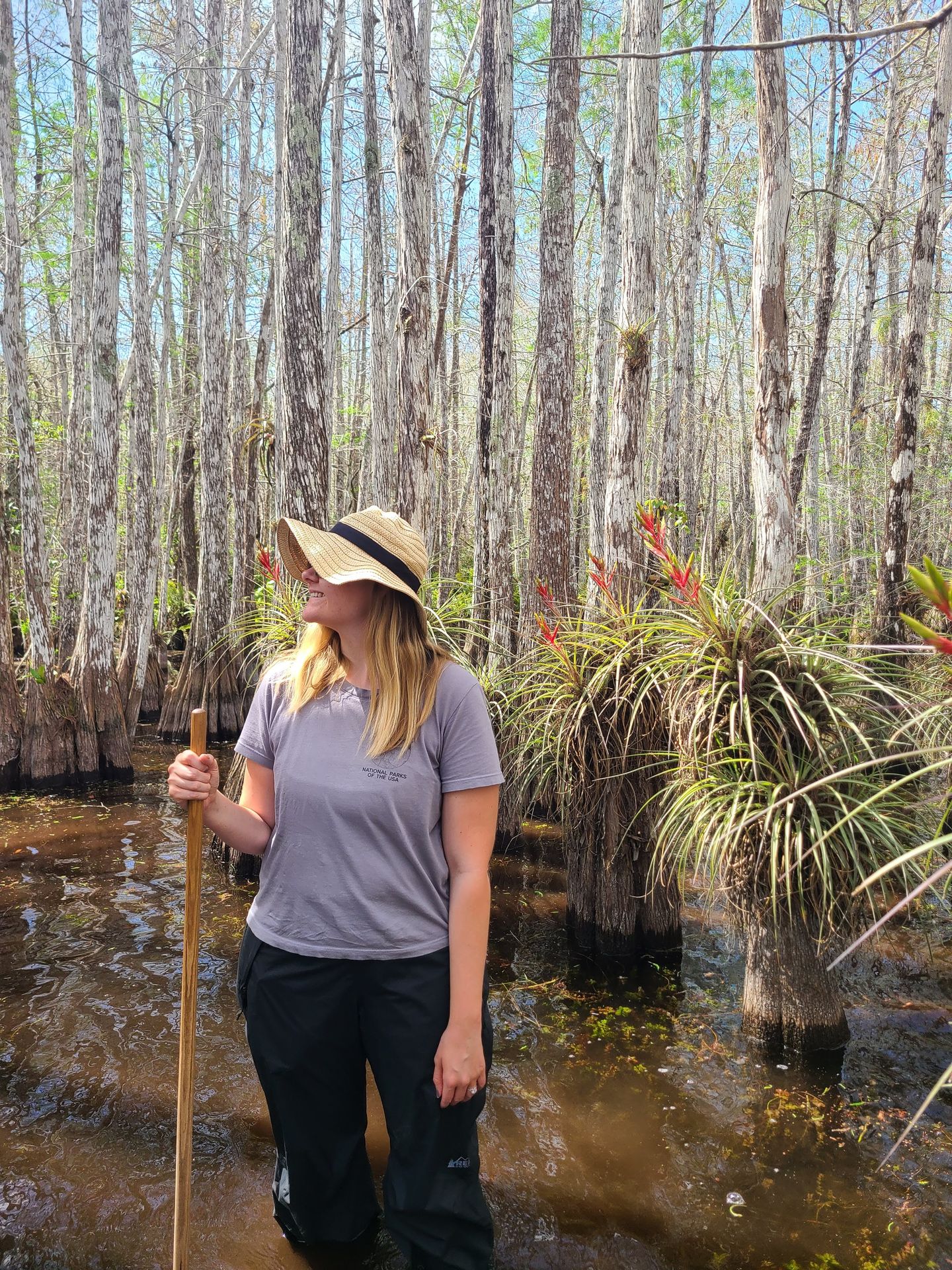 Lydia standing knee deep in water among cypress trees. A orange flower blooms out of greenery attached to a tree behind her